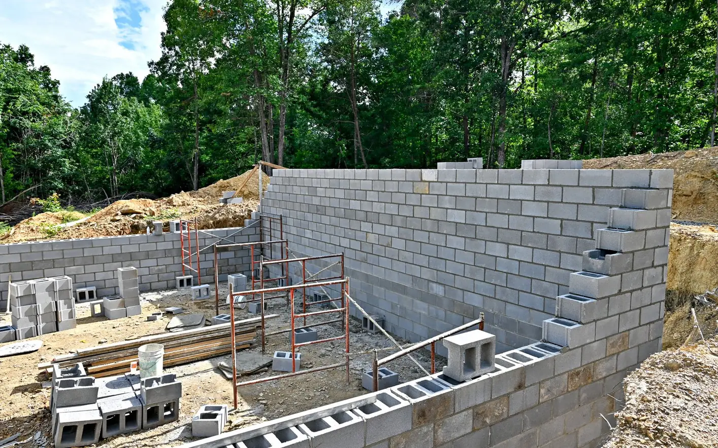 Concrete block basement walls rising on a wooded lot with scaffolding and stacked block, block foundations