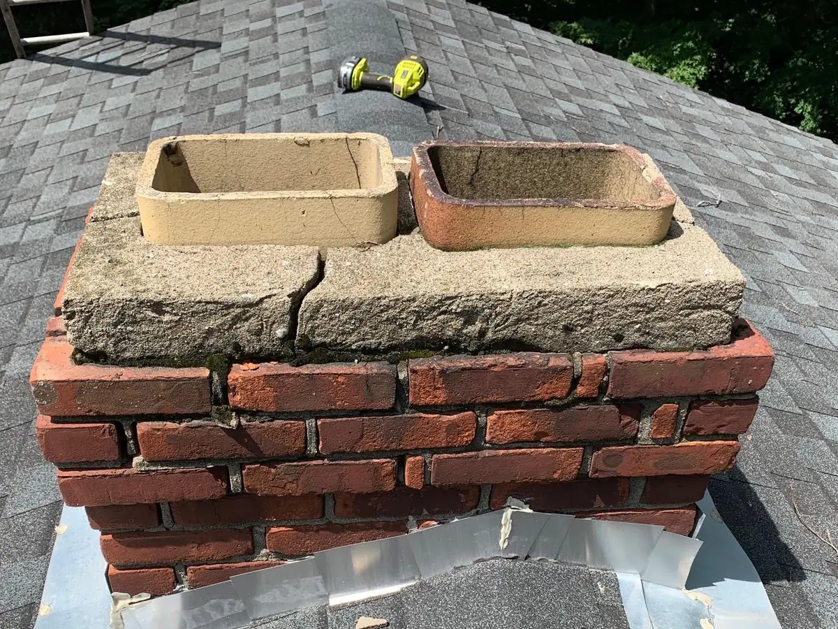 Short brick chimney on a shingle roof with a cracked concrete crown around two clay flue tiles, chimney repair