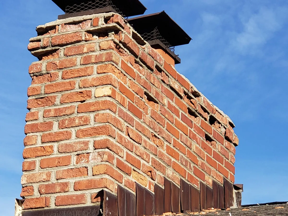 Brick chimney above a roofline with loose, crumbling top courses and open mortar joints, chimney repair